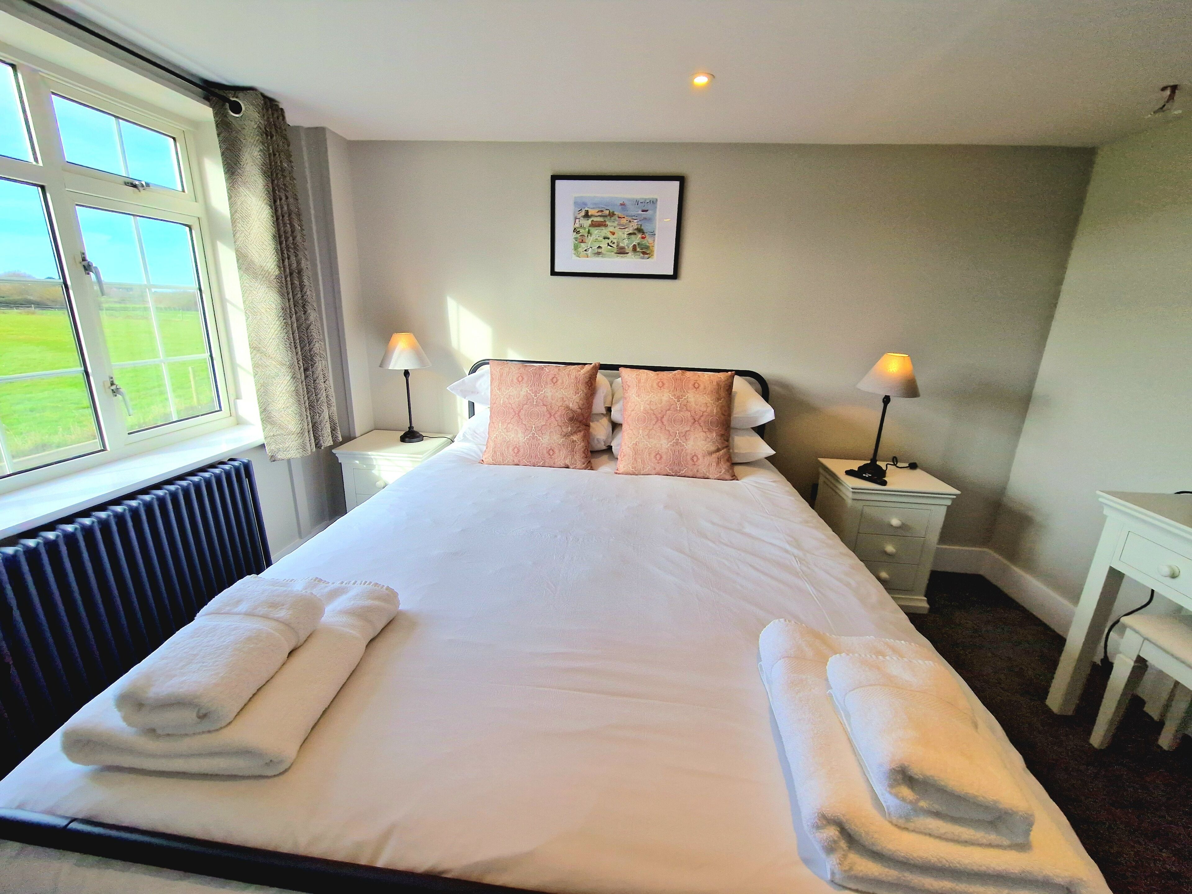 A neatly made bed with pillows and folded towels, a bedside table with a lamp, a desk, and a window with curtains.