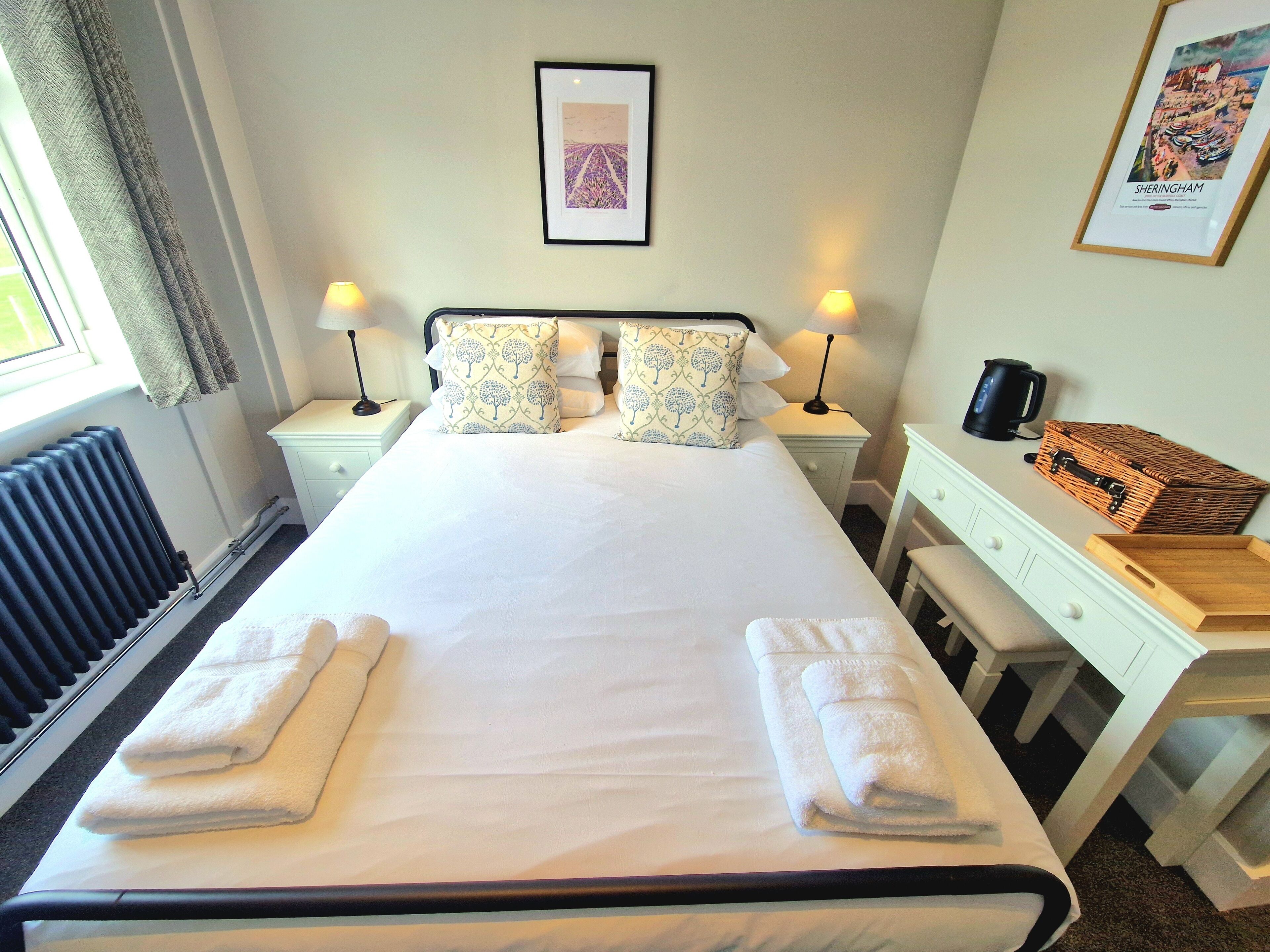 A neatly made bed with pillows, a bedside table with a lamp, a framed picture on the wall, and a wicker basket on the nightstand.