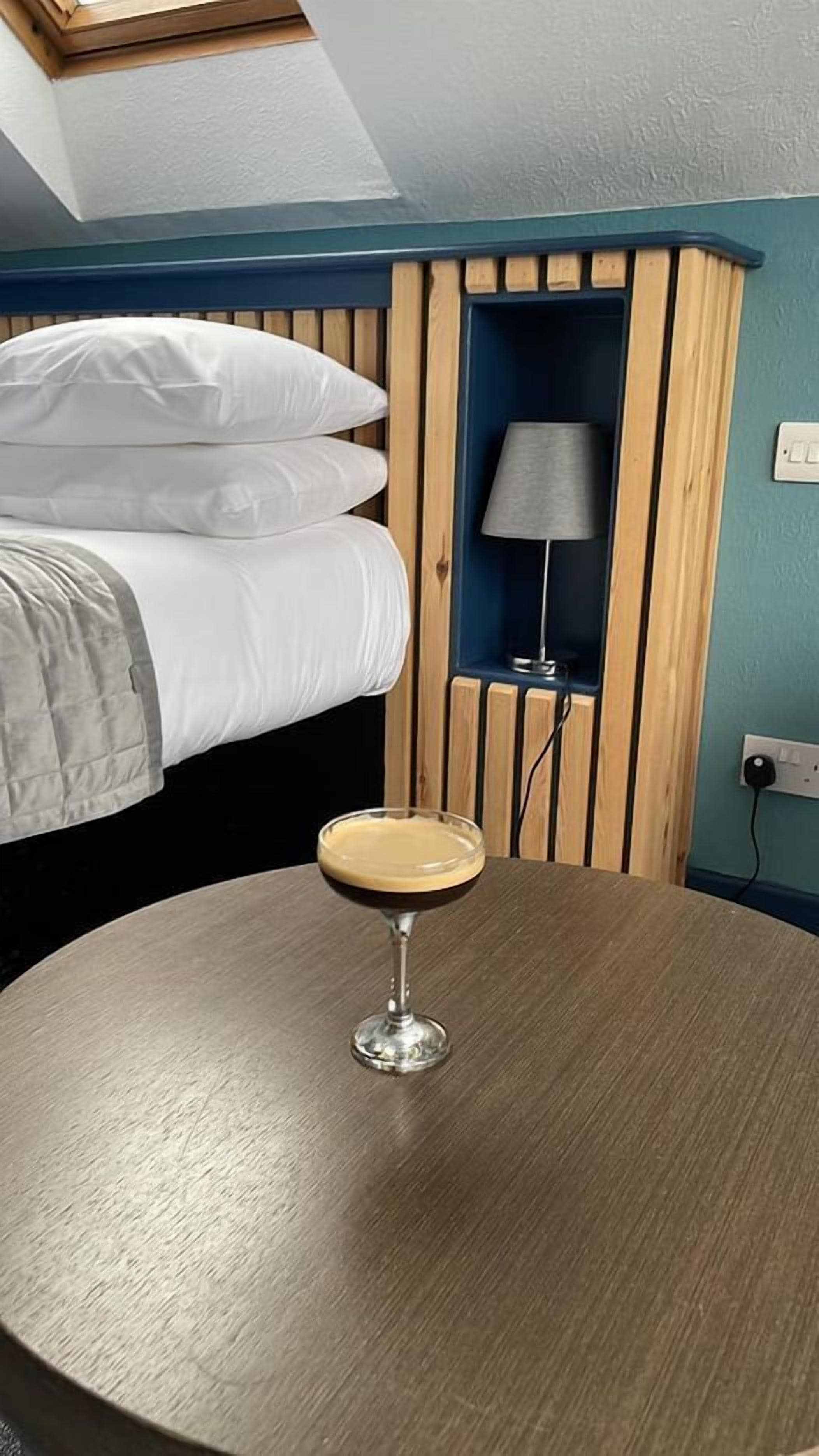 Una habitación con una cama, una mesa redonda con una bebida, una lámpara y un estante fijado a la pared.