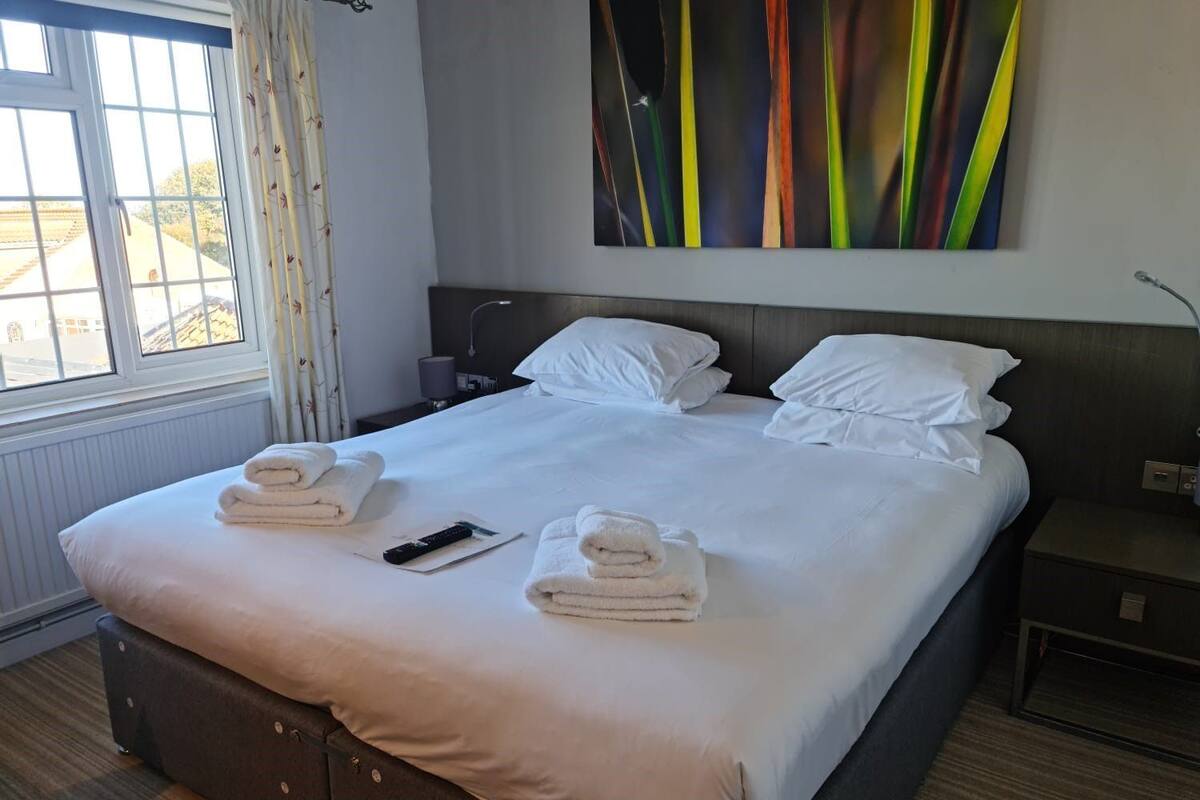 A hotel room with a large bed, two bedside tables, a window with curtains, a painting on the wall, and a remote control on the bed.