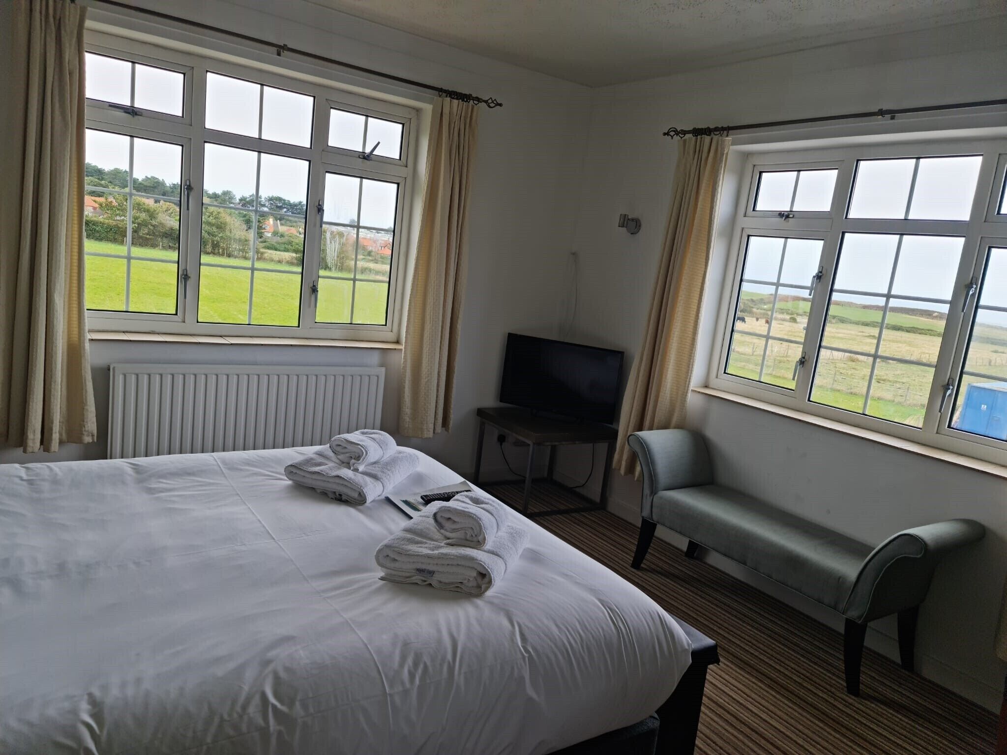 Een hotelkamer met een bed, een televisie, een bankje en grote ramen met uitzicht op de omgeving.