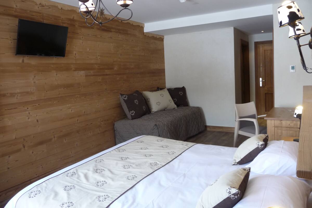 A bedroom with a wooden wall, a bed with patterned bedding, a desk with a lamp, and a chair.
