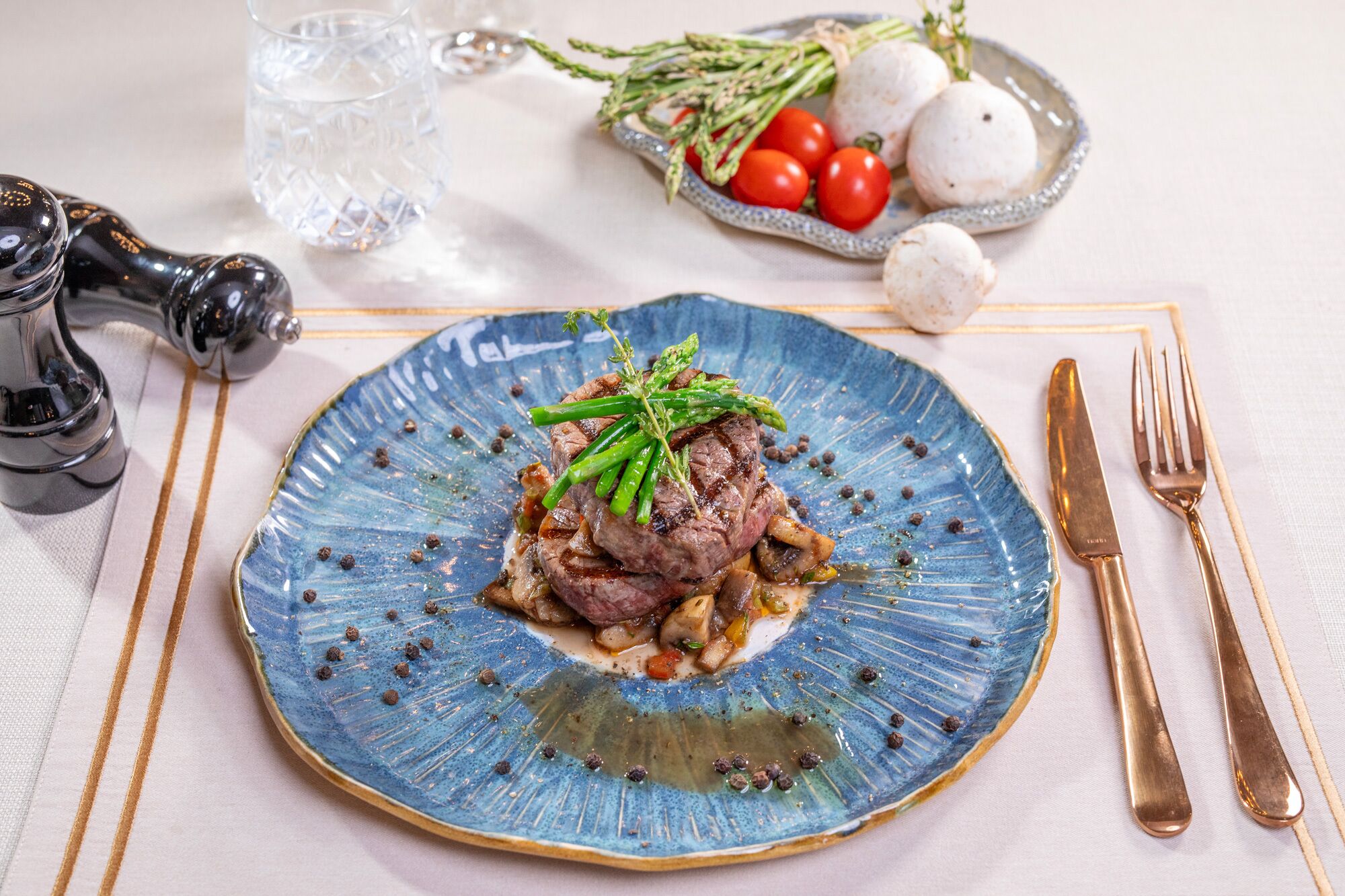 A blue plate with a steak topped with asparagus, mushrooms, and a sauce, accompanied by a glass of water, a salt and pepper shaker, and a small dish with tomatoes and herbs.