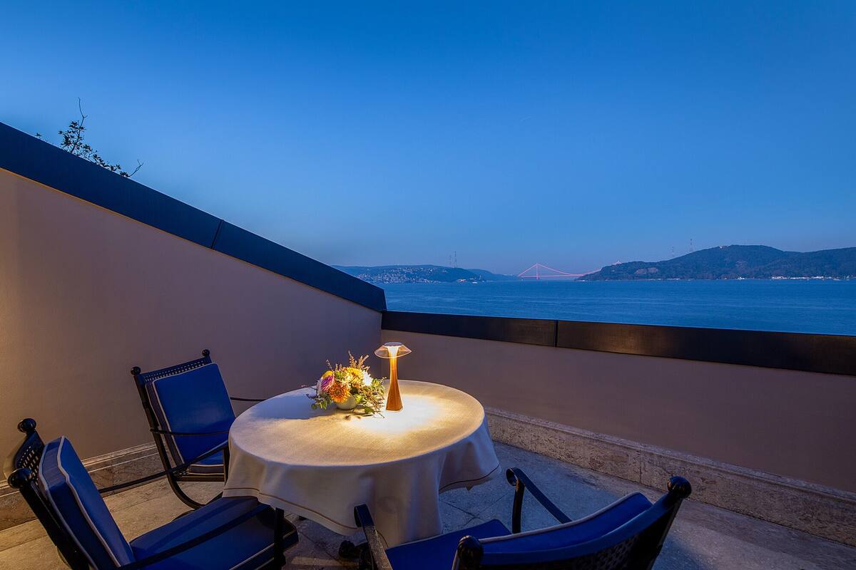 A round table with a white tablecloth and two blue chairs on a balcony overlooking a body of water with a bridge in the distance.