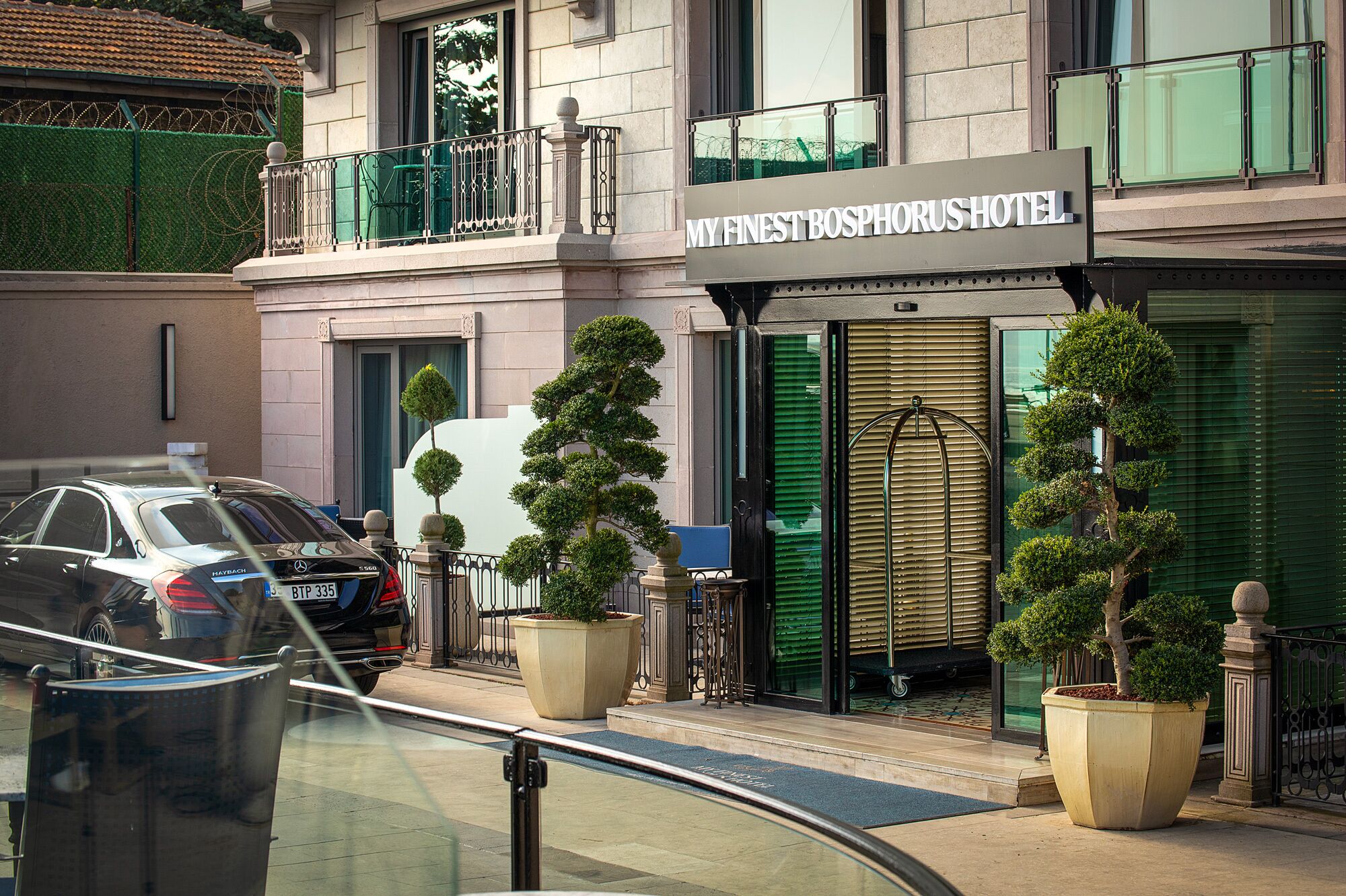 Entrada de um hotel com uma placa que diz "MY FINEST BOSPHORUS HOTEL". Há duas árvores em vasos na frente da entrada, e um carro estacionado na rua.