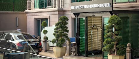Entrada de um hotel com uma placa que diz "MY FINEST BOSPHORUS HOTEL". Há duas árvores em vasos na frente da entrada, e um carro estacionado na rua.