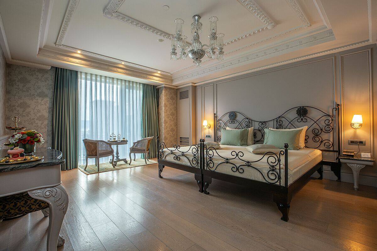 A hotel room with a large bed, a desk with a chair, a chandelier, and a view of the outdoors through a window with curtains.