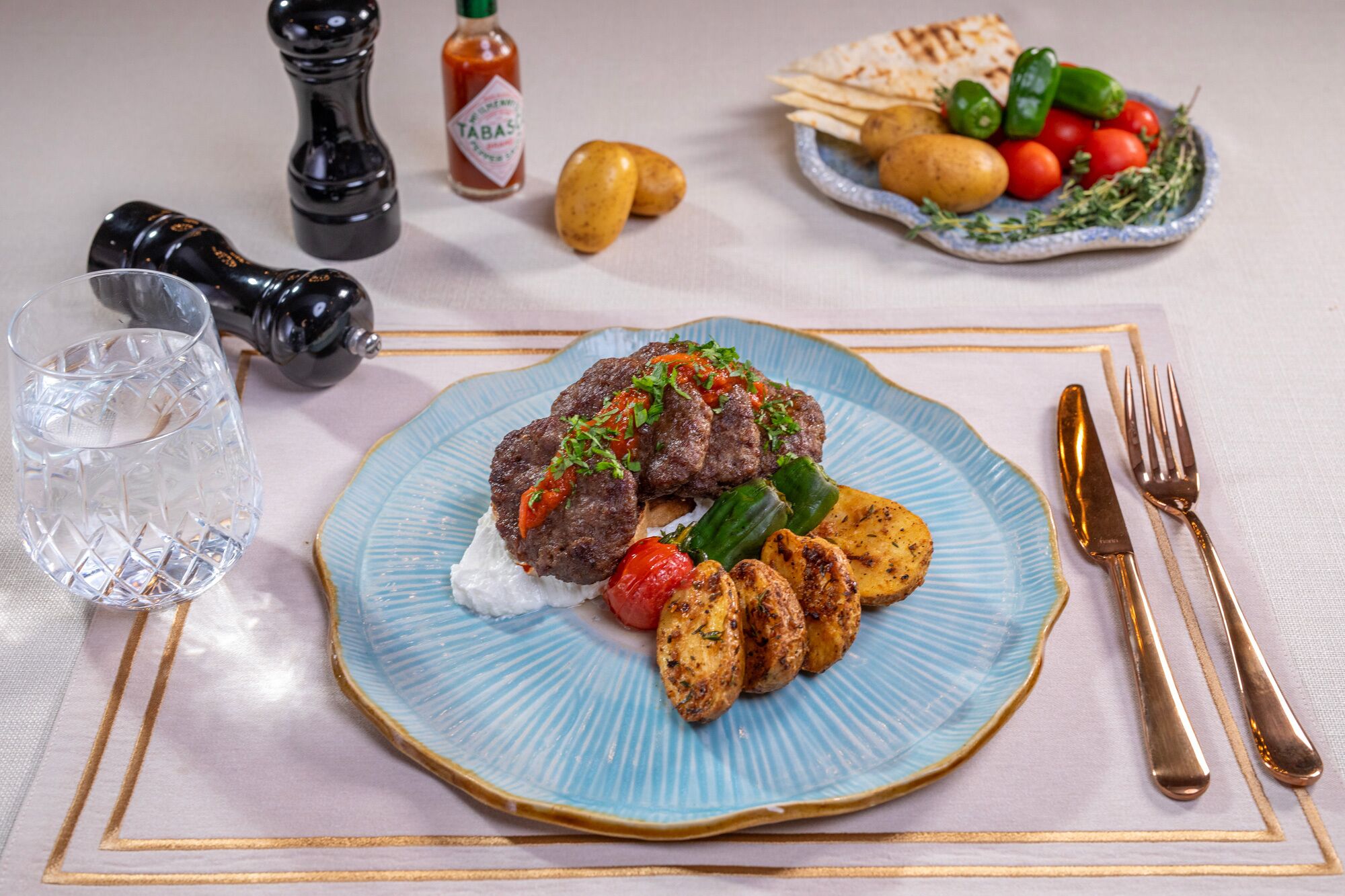 A plate of food with a blue rim, a glass of water, and a bottle of Tabasco sauce on a table.