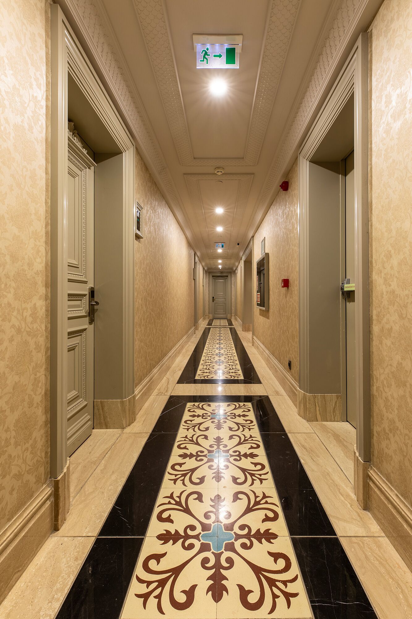 Un largo pasillo de hotel, estrecho, con paredes con diseño y suelo de baldosas negras y beige.