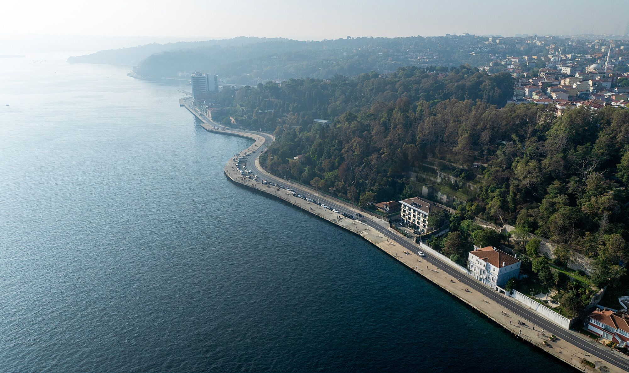 An aerial view of a coastal city with a long, curved road along the shoreline, dense forested hills in the background, and a prominent building with a dome structure.