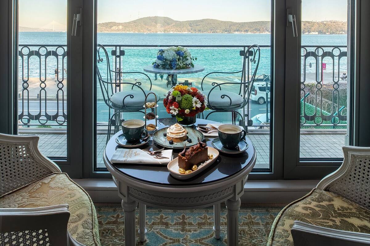 A room with a large window overlooking a body of water and a bridge. There is a table with a bouquet of flowers, a plate of food, and two cups of coffee.