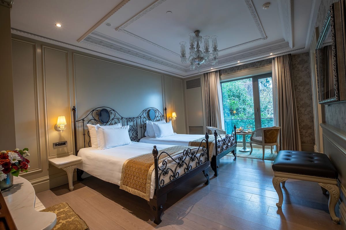 A hotel room with two beds, a chandelier, a balcony with a chair, and a view of greenery.