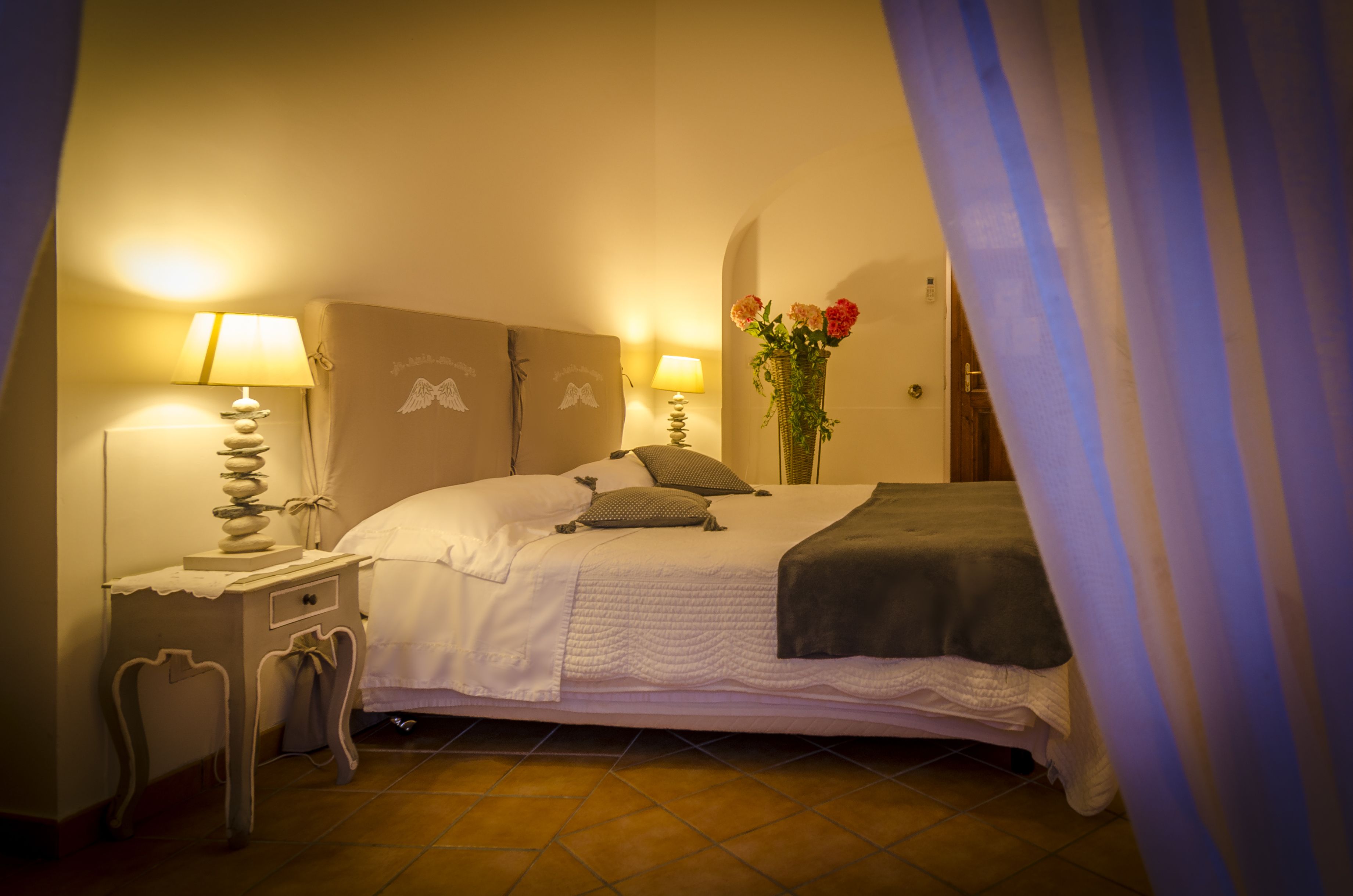 A bedroom with a bed, two bedside lamps, a nightstand, a vase with flowers, and a curtain.