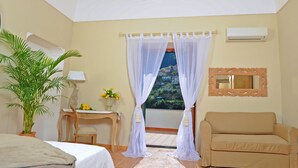Un dormitorio con cama, escritorio, silla, sofá, planta en maceta y ventana con cortinas translúcidas.