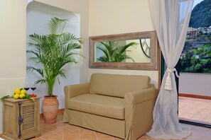 A beige sofa, a potted plant, a small wooden cabinet, and a mirror on a balcony with a view of a hillside. - Maison Liparlati Positano (Positano)