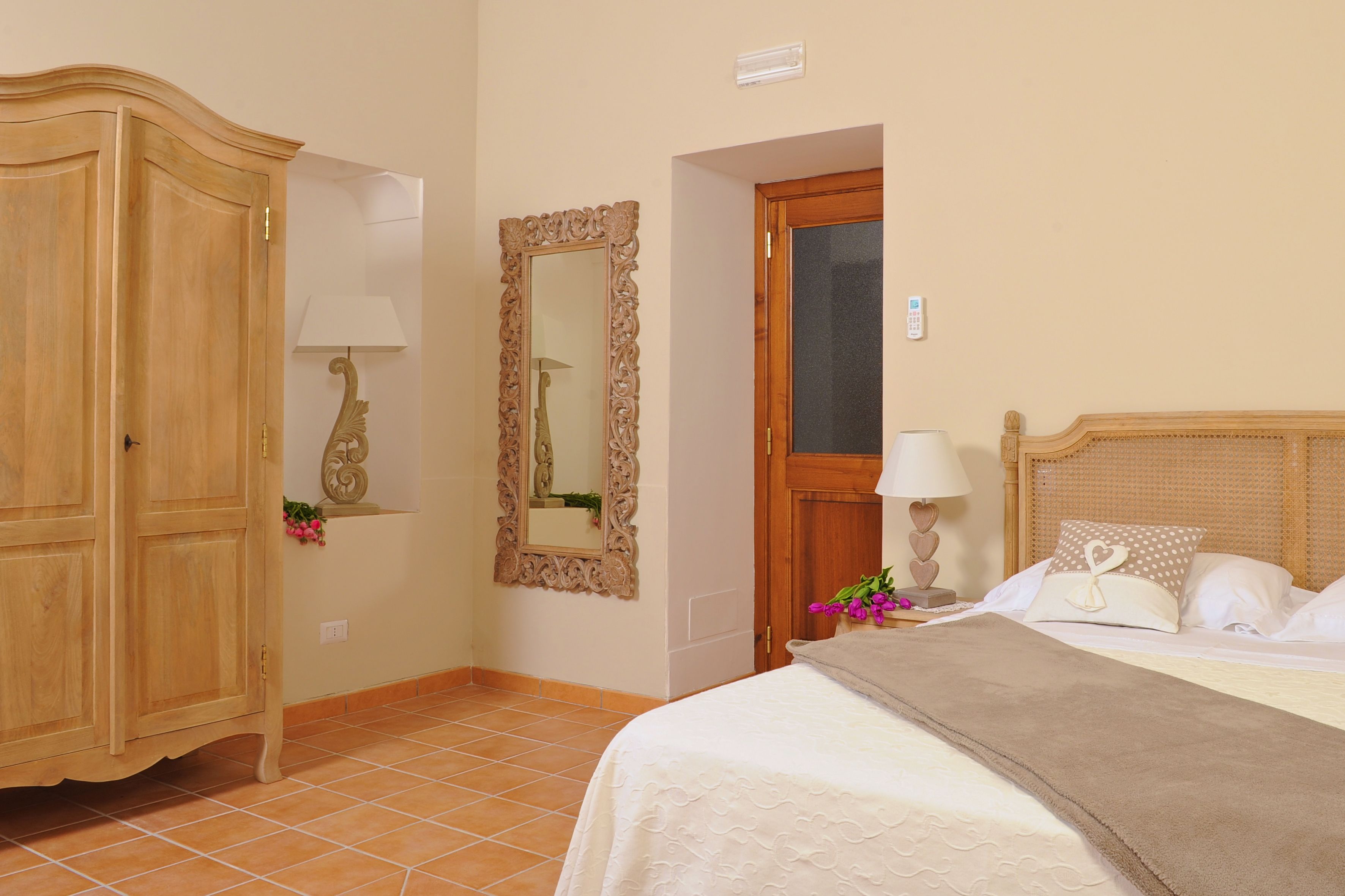 A bedroom with a wooden wardrobe, a mirror, a bed with white bedding, and a nightstand with a lamp.