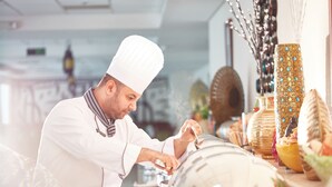 Ein Koch in weißer Uniform bereitet Essen in einer Küche mit dekorativen Vasen und modernem Design zu.