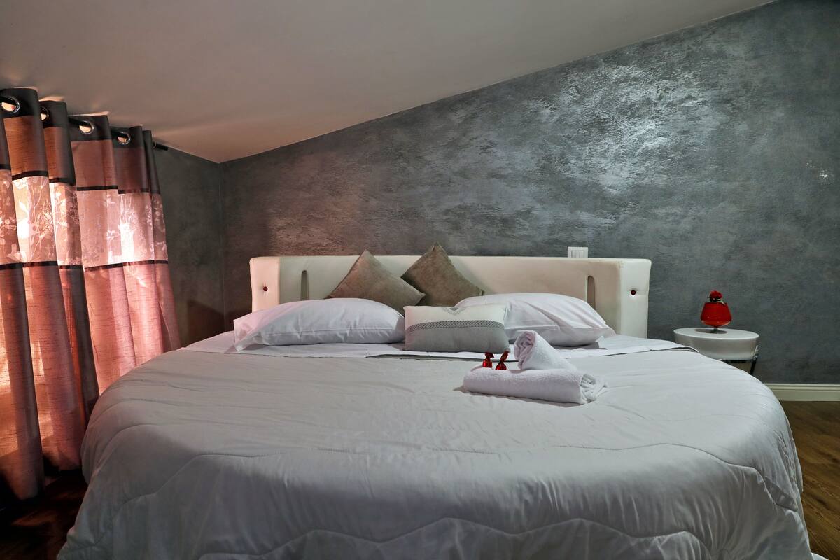 Un lit bien fait, avec une literie blanche, une tête de lit agrémentée de plusieurs coussins et une petite table de chevet sur laquelle se trouve un objet rouge.