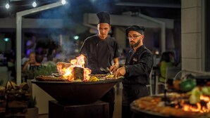 Des chefs préparent des plats sur un grand grill, avec une foule en arrière-plan.