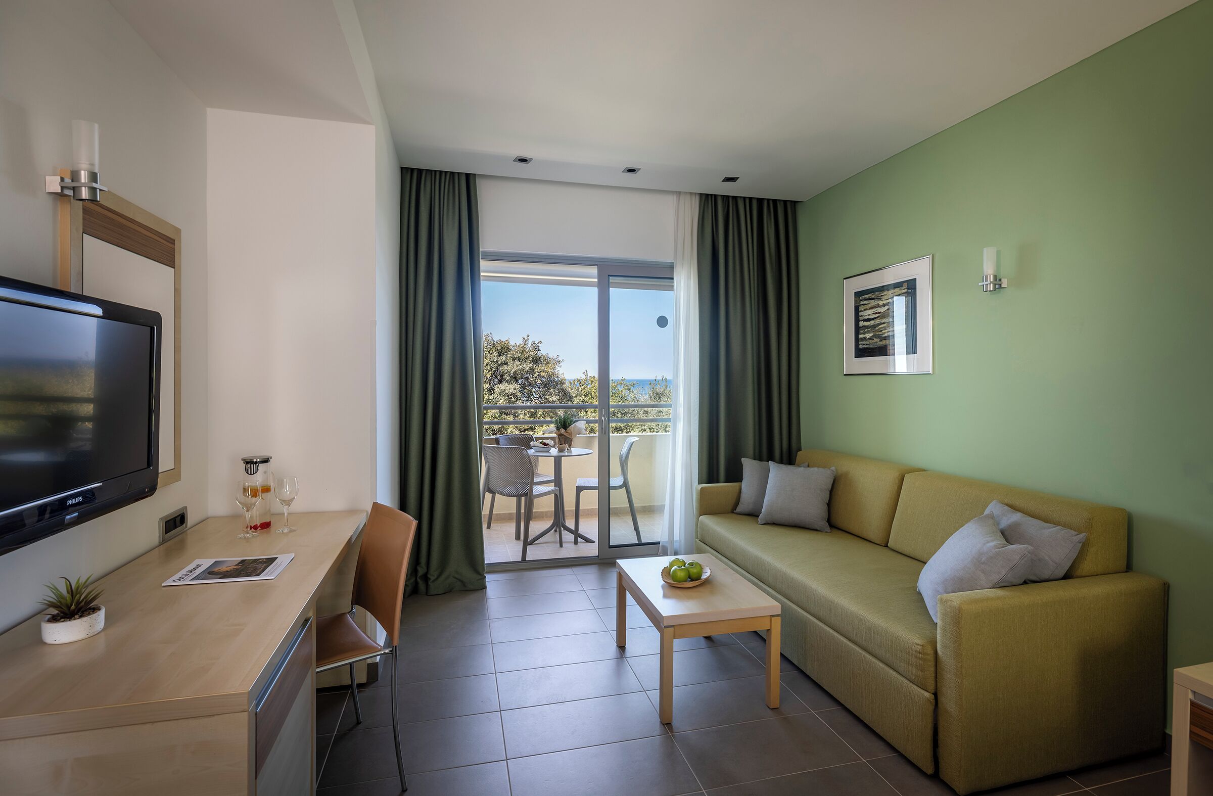 A modern hotel room with a sofa, a TV, a dining table, and a balcony with a view.