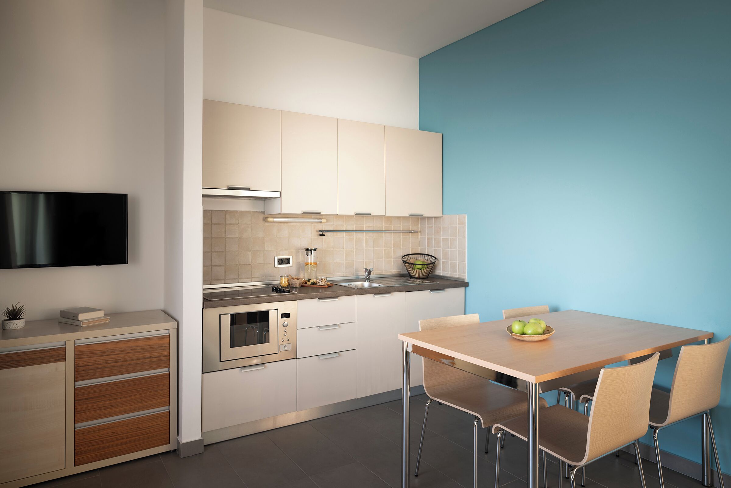 Una cocina moderna con armarios blancos, horno empotrado, microondas y un lavabo. Hay una mesa de comedor de madera con dos sillas.