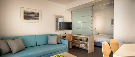A modern hotel room with a sofa, a dining table, a TV, and a bed with a sliding door.