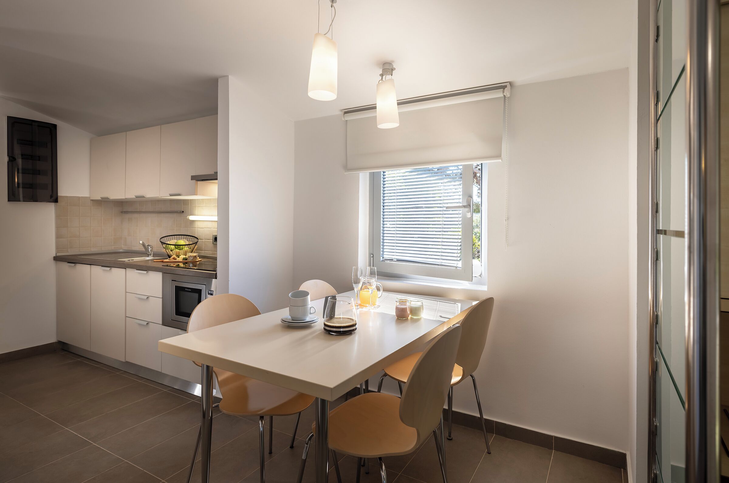 Egy modern konyha fehér szekrényekkel, egy étkezőasztallal, hozzáillő székekkel, ablakon elhelyezett reluxával és mennyezetről lógó lámpákkal.