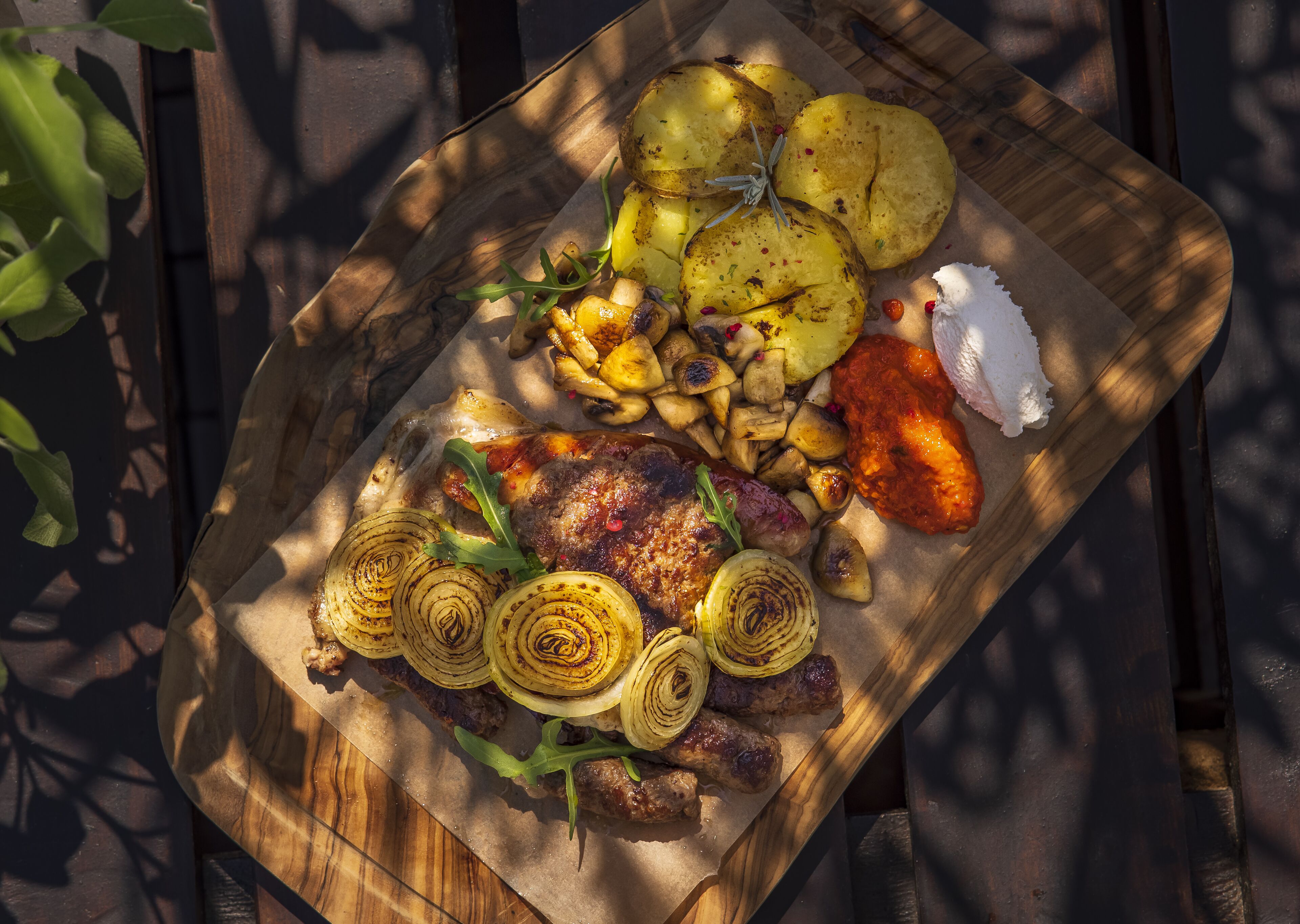Una tabla de madera con carne a la parrilla, patatas asadas, cebollas a la brasa y una guarnición de salsa.
