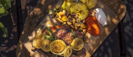Une planche en bois servant de plat, sur laquelle on présente de la viande grillée, des pommes de terre rôties, des oignons grillés et une sauce en accompagnement.