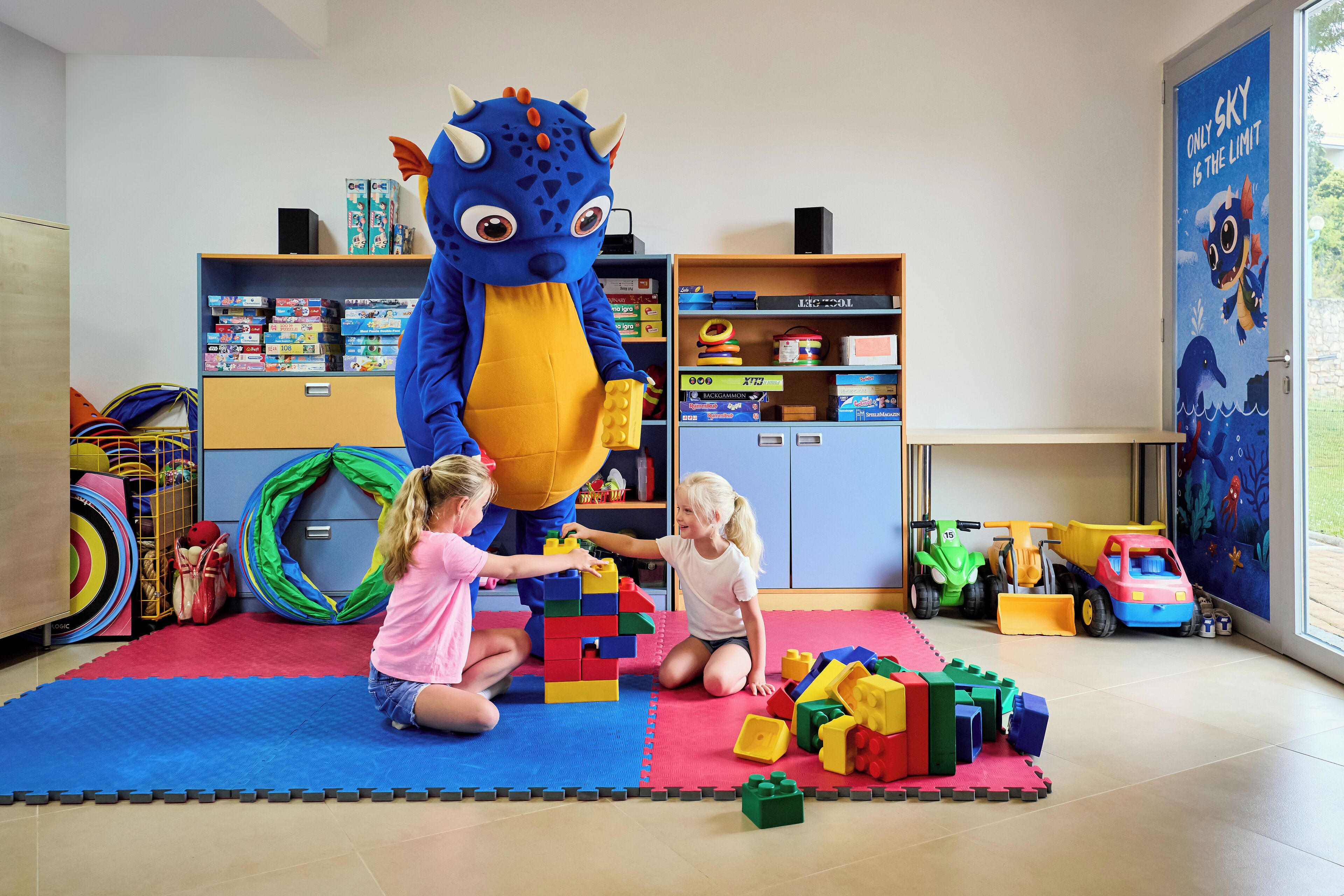 A playroom with a large blue dinosaur mascot, children playing with building blocks, and a window with a view of the outdoors.
