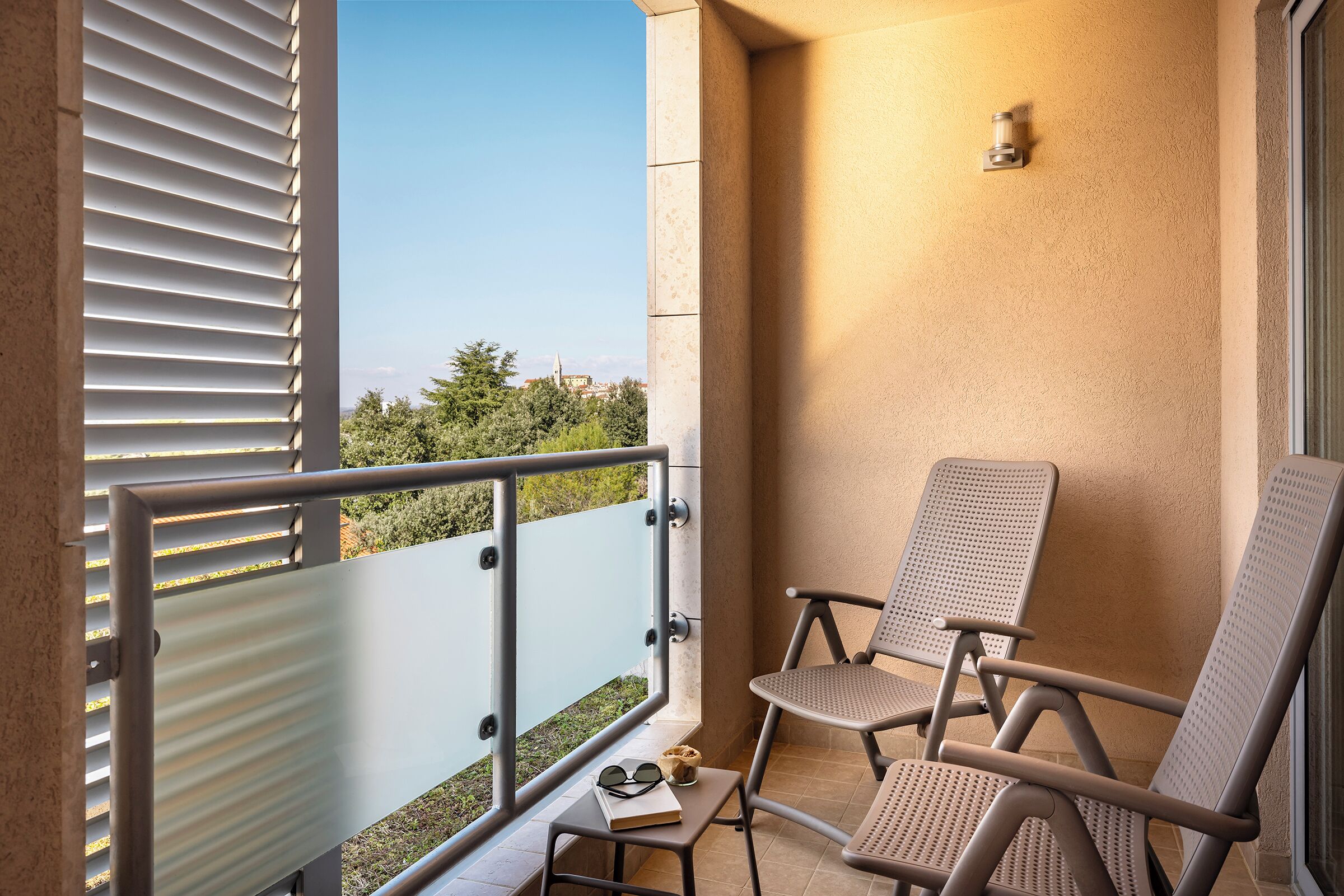 A balcony with two chairs, a small table with a book and sunglasses, and a view of greenery and a distant building.