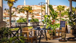 Un café en plein air avec des tables et des chaises, entouré de plantes en pot et offrant une vue sur un paysage urbain avec des bâtiments et des palmiers.