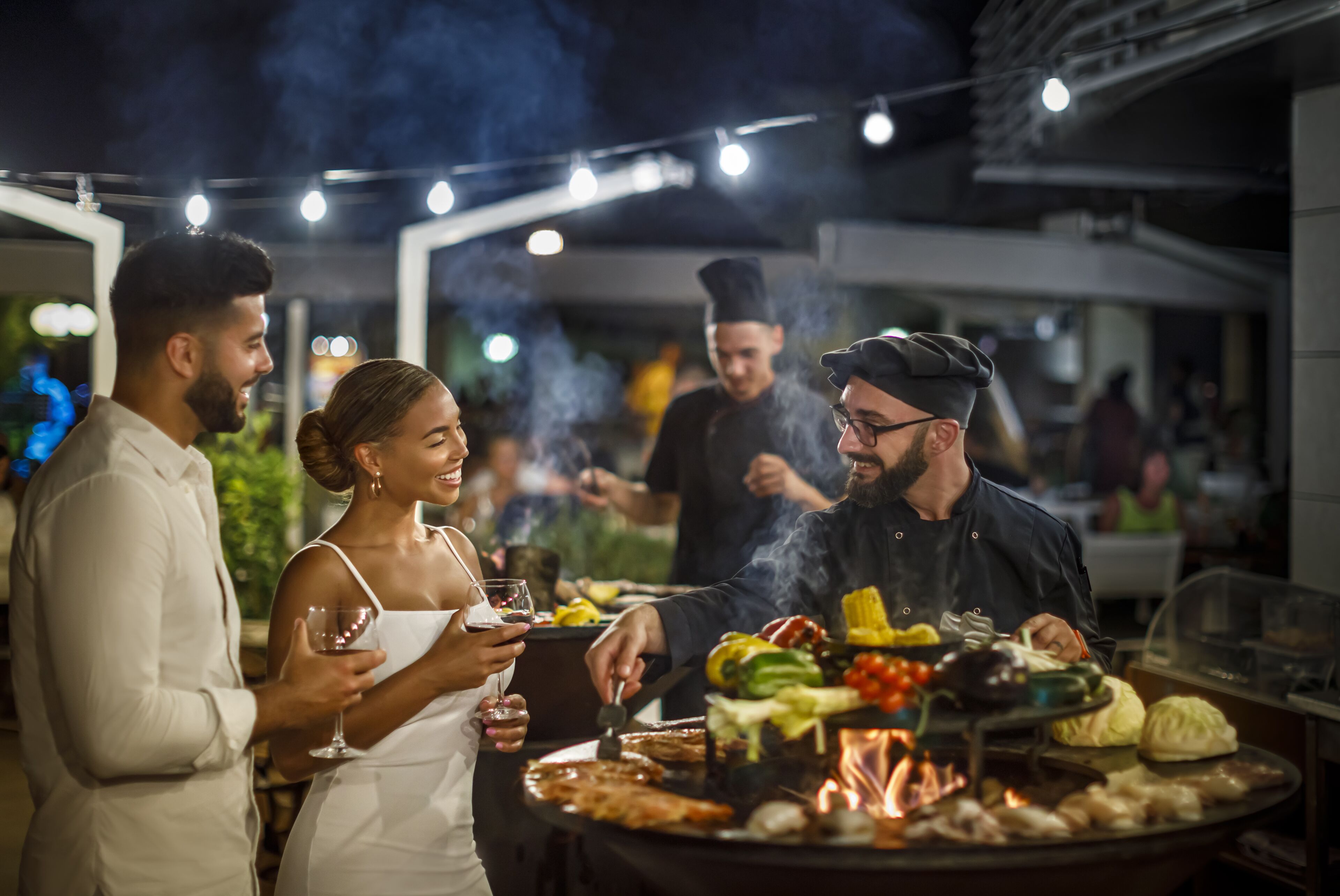 A group of people dining outdoors at night with a grill and various foods.