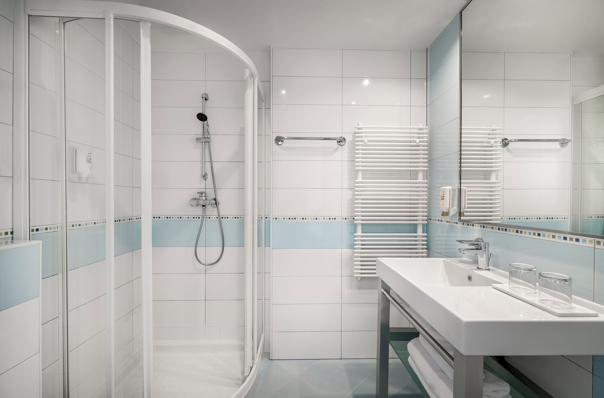 A modern bathroom with a glass shower enclosure, a white sink with two glasses, and a towel rack.