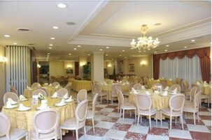 Un salón de banquetes con mesas redondas dispuestas para cenar, sillas blancas y una araña de cristal.