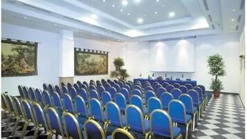 Un salón de conferencias con filas de sillas azules, un escenario y dos cuadros en las paredes.