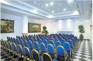 Una sala conferenze con file di sedie blu, un palco e due quadri alle pareti.
