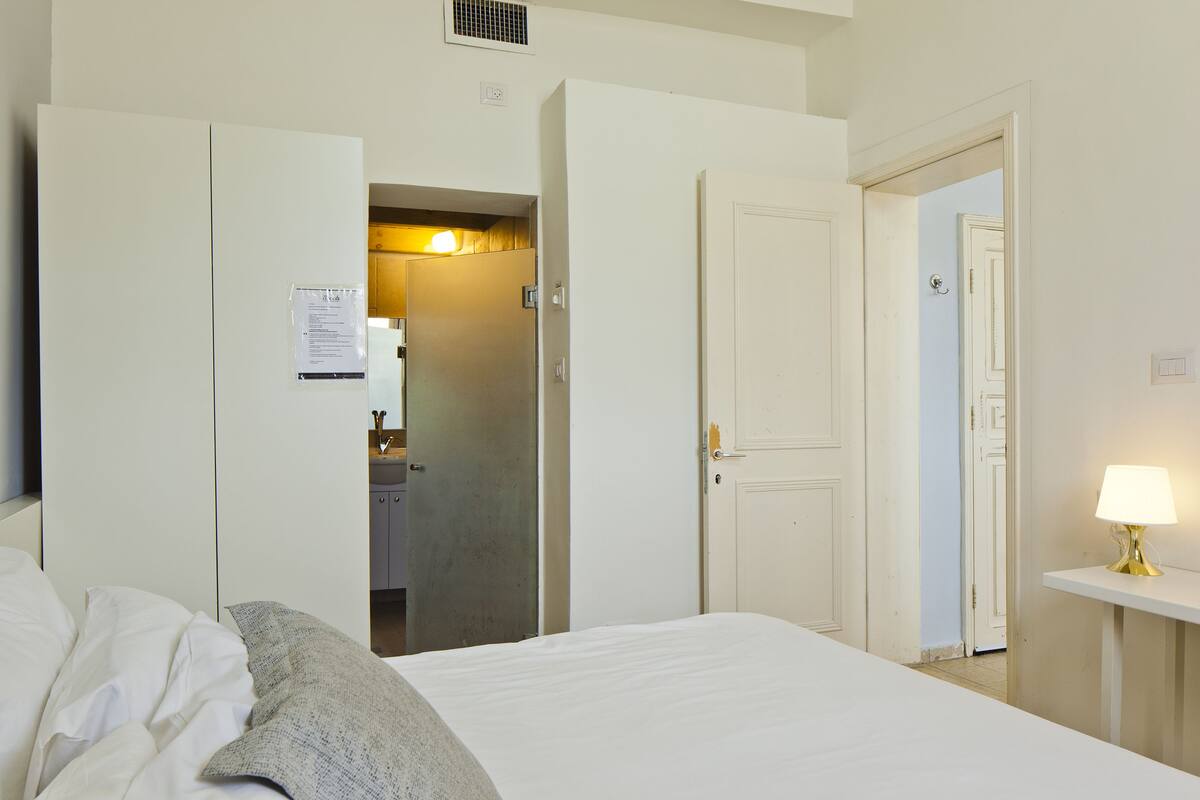 A hotel room with a bed, a bedside table with a lamp, a wardrobe, and an open door leading to a bathroom.