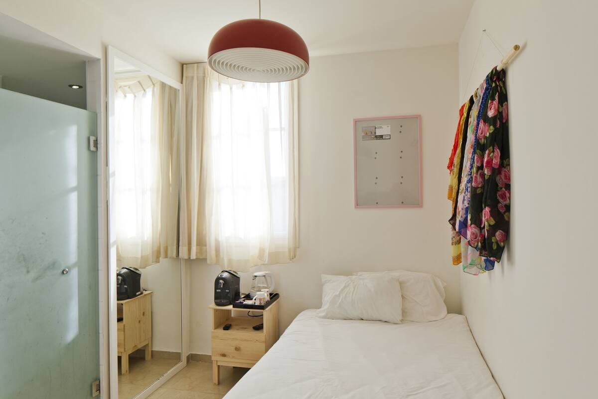A bedroom with a bed, a nightstand with a coffee maker, a wardrobe, and a window with curtains.