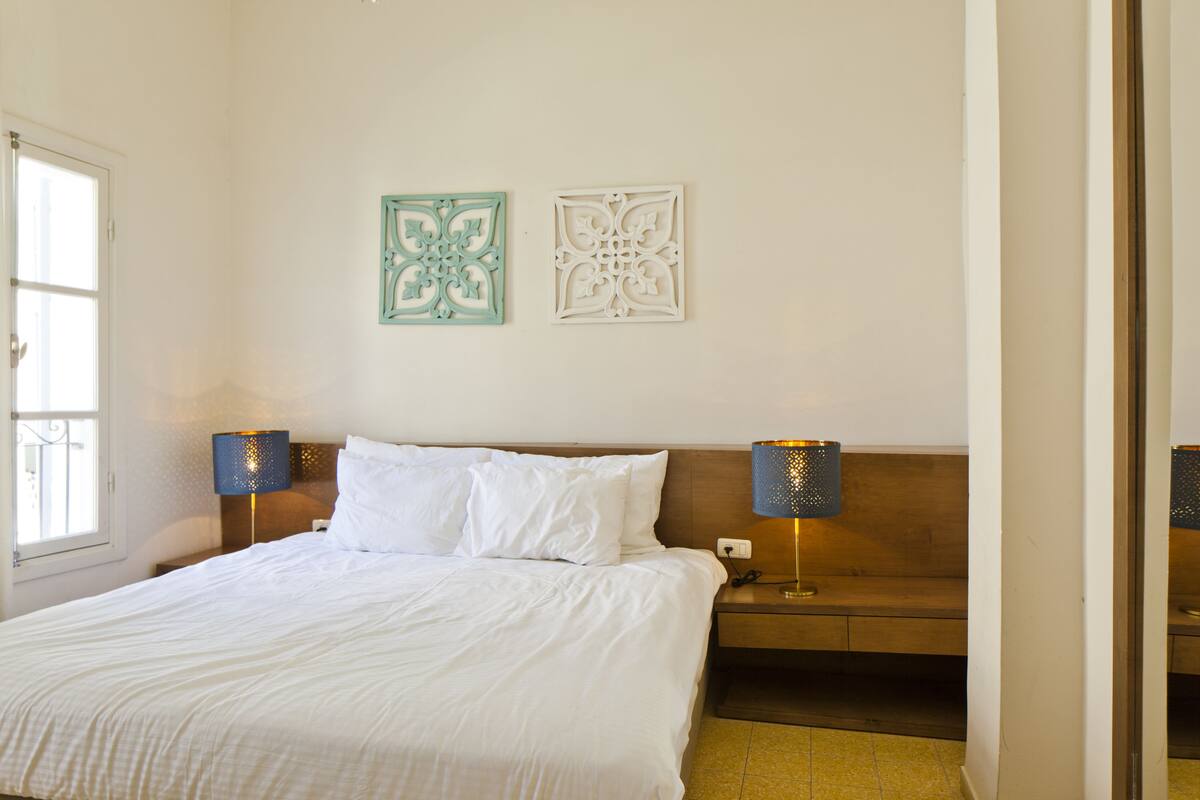 A neatly made bed with white linens, two wall-mounted lamps, and two decorative wall pieces.
