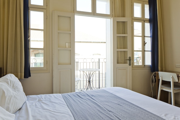 A hotel room with a bed, a small table, and a chair. There is a balcony with a railing and a view of the outside.