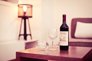 A bottle of wine and two glasses on a wooden table in a room with a lamp and a bed in the background.