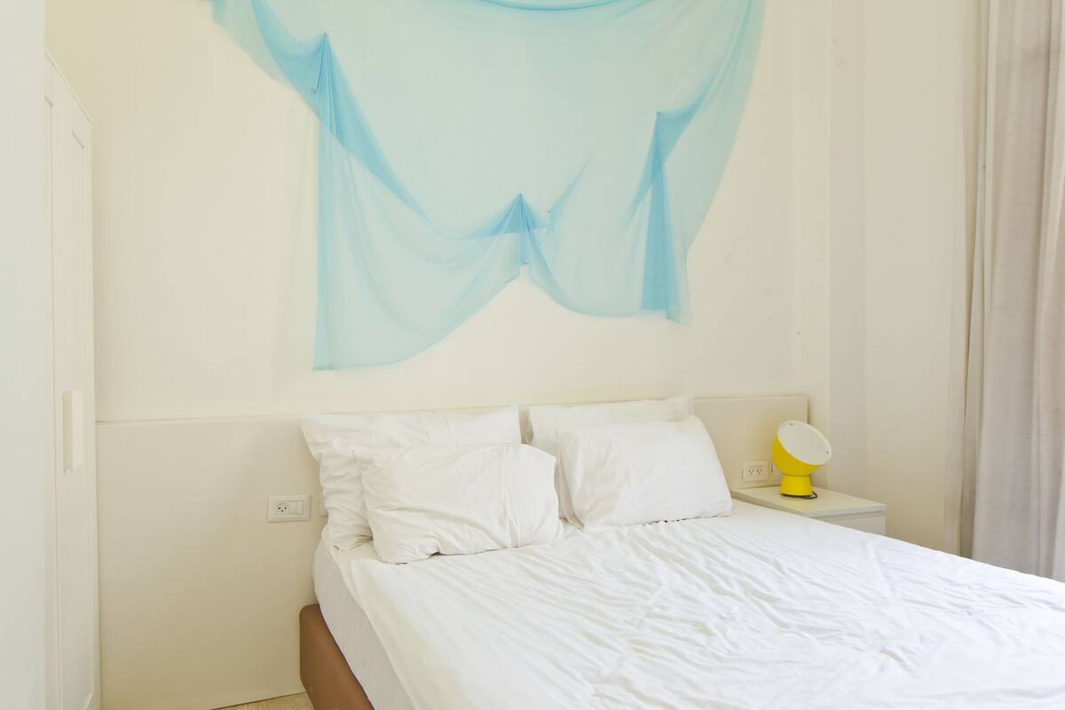 A bedroom with a bed, white pillows, a yellow bedside lamp, and a blue sheer curtain.