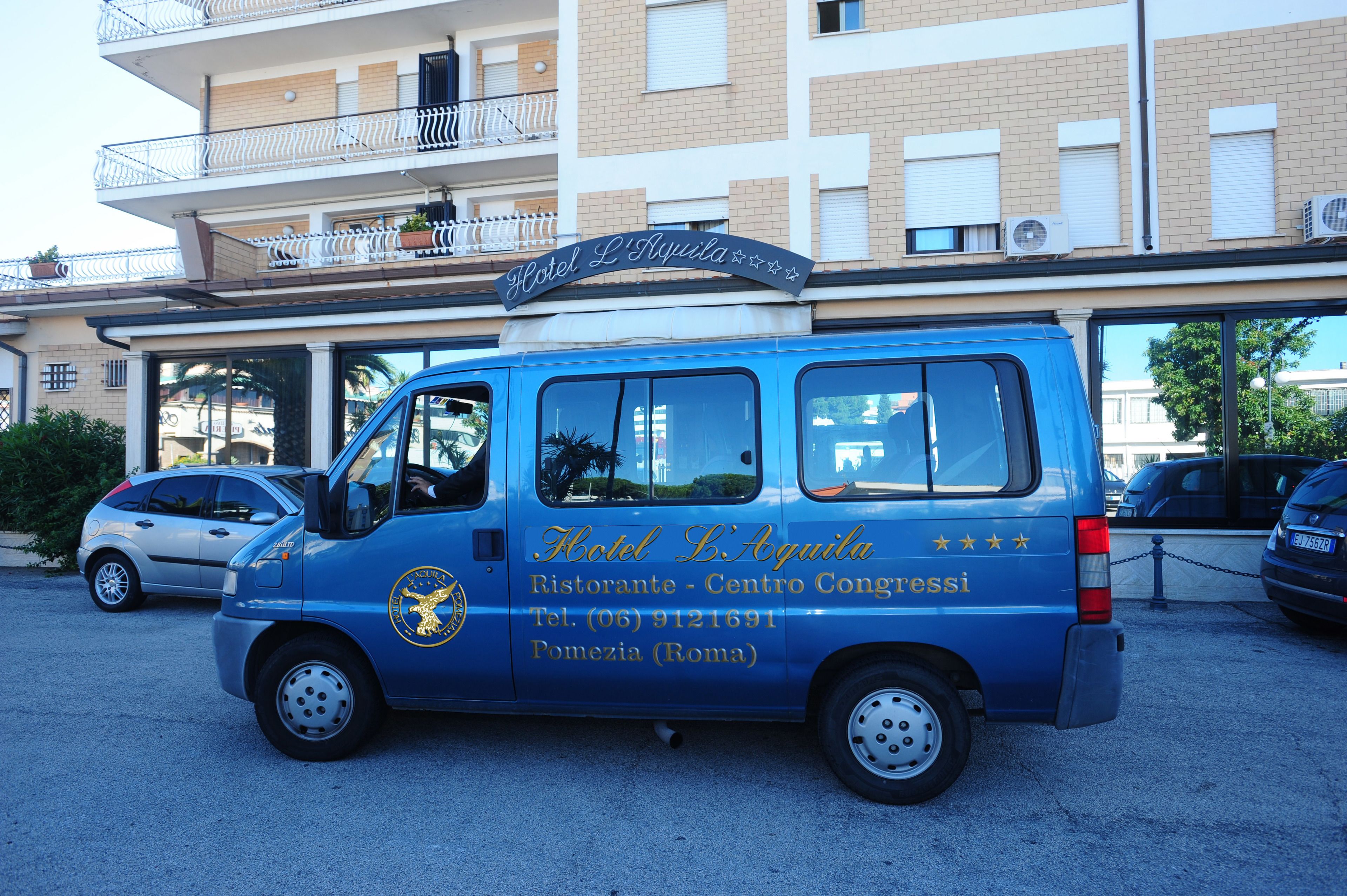 A blue van with the sign "Hotel L'Aquila Restaurante - Centro Congressi" and contact information.