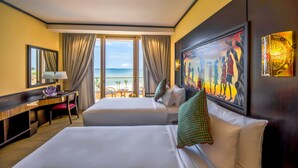 Habitación de hotel con dos camas, un escritorio, una silla y un ventanal con vista a la playa.