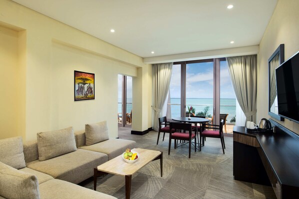 A modern hotel room with a sofa, dining table, and a TV. The room has large windows with a view of the sea.