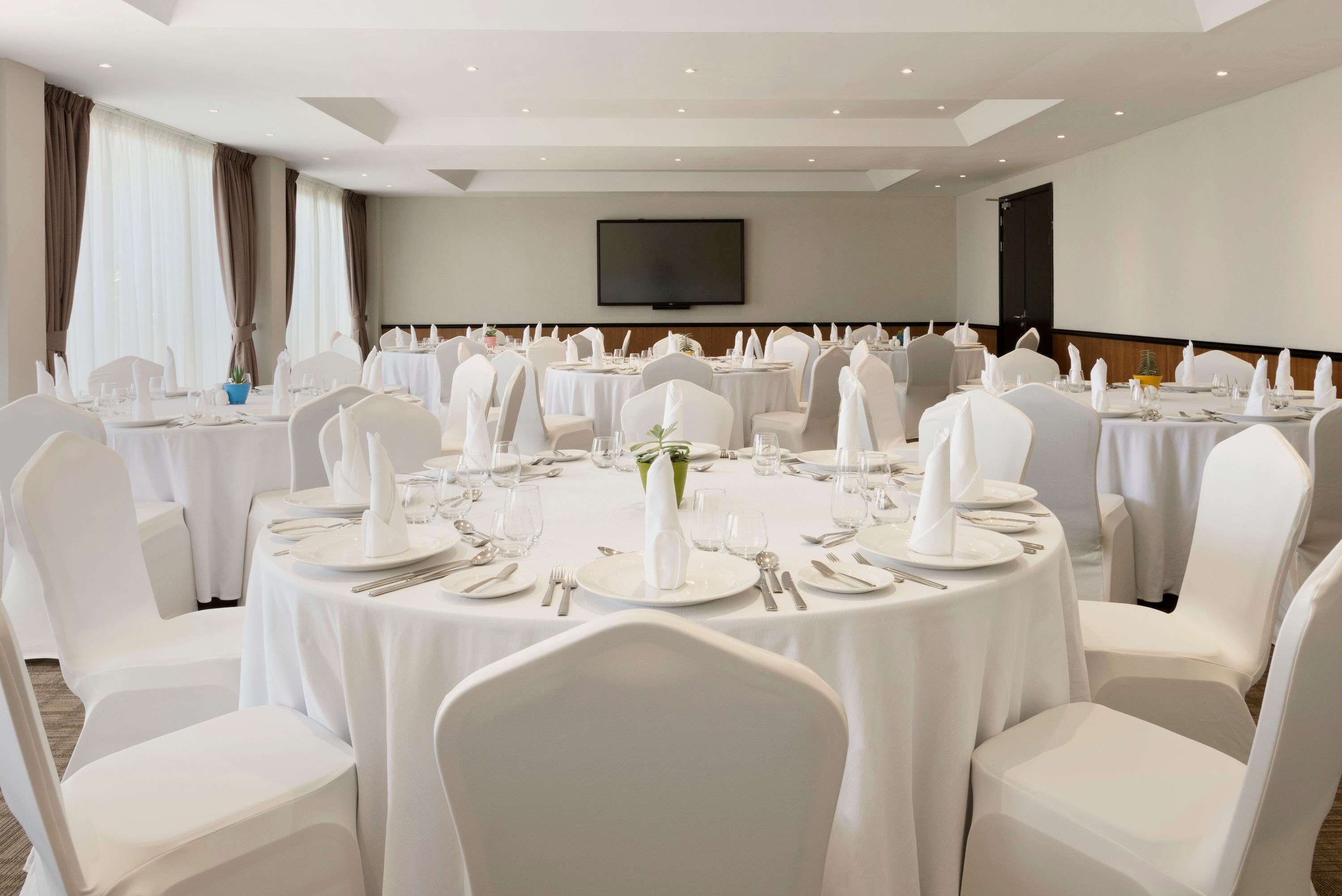 A banquet hall with round tables set for dining, white chairs, and a flat-screen TV mounted on the wall.