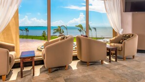 Una habitación con un ventanal grande con vistas a la playa, dos sillones, una mesita y un televisor de pantalla plana montado en la pared.