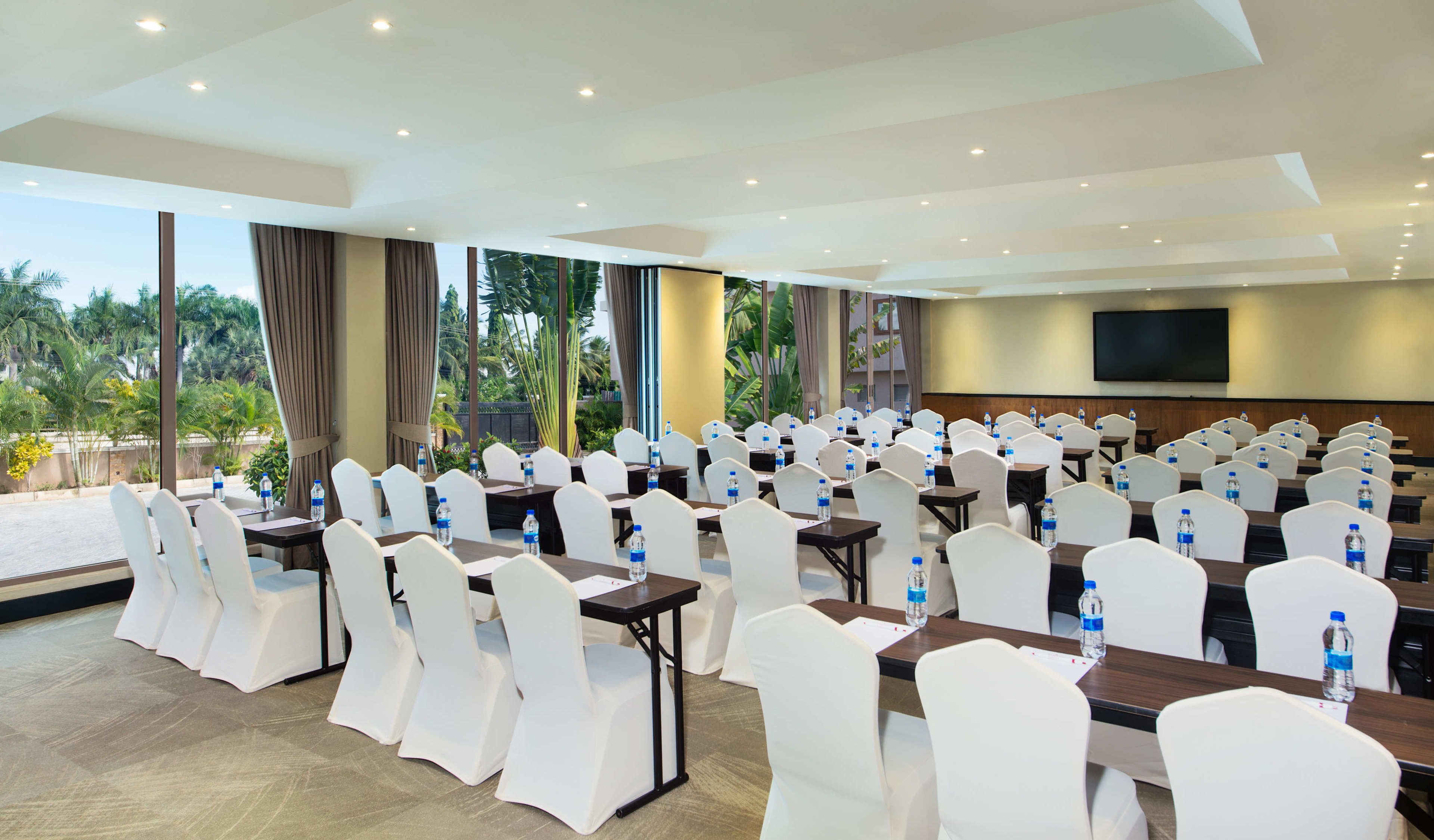 Un salón de conferencias con filas de sillas y mesas blancas, botellas de agua y una pantalla grande.