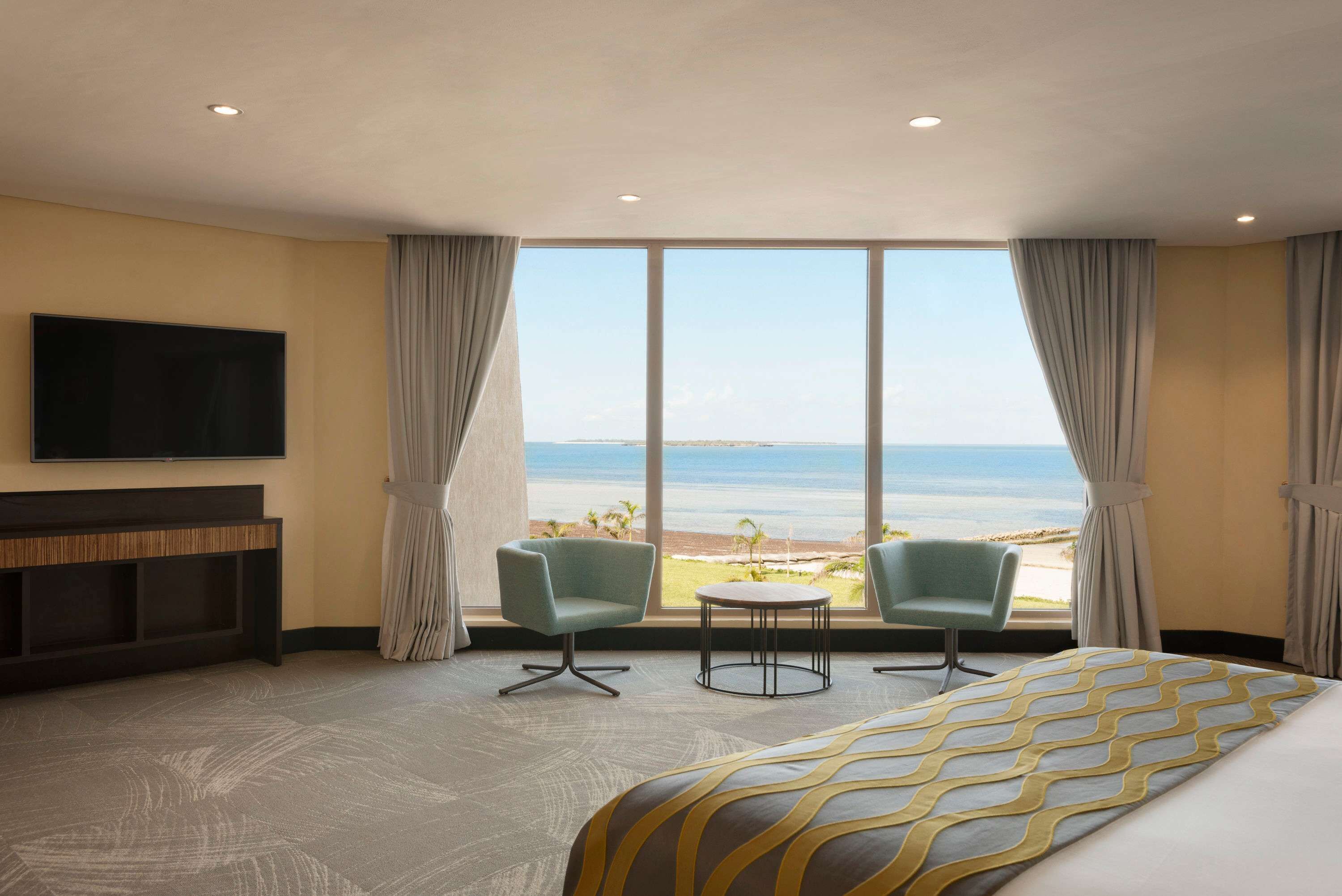 A hotel room with a large window offering a view of the beach, a bed with a patterned bedspread, a television mounted on the wall, and two chairs near the window.